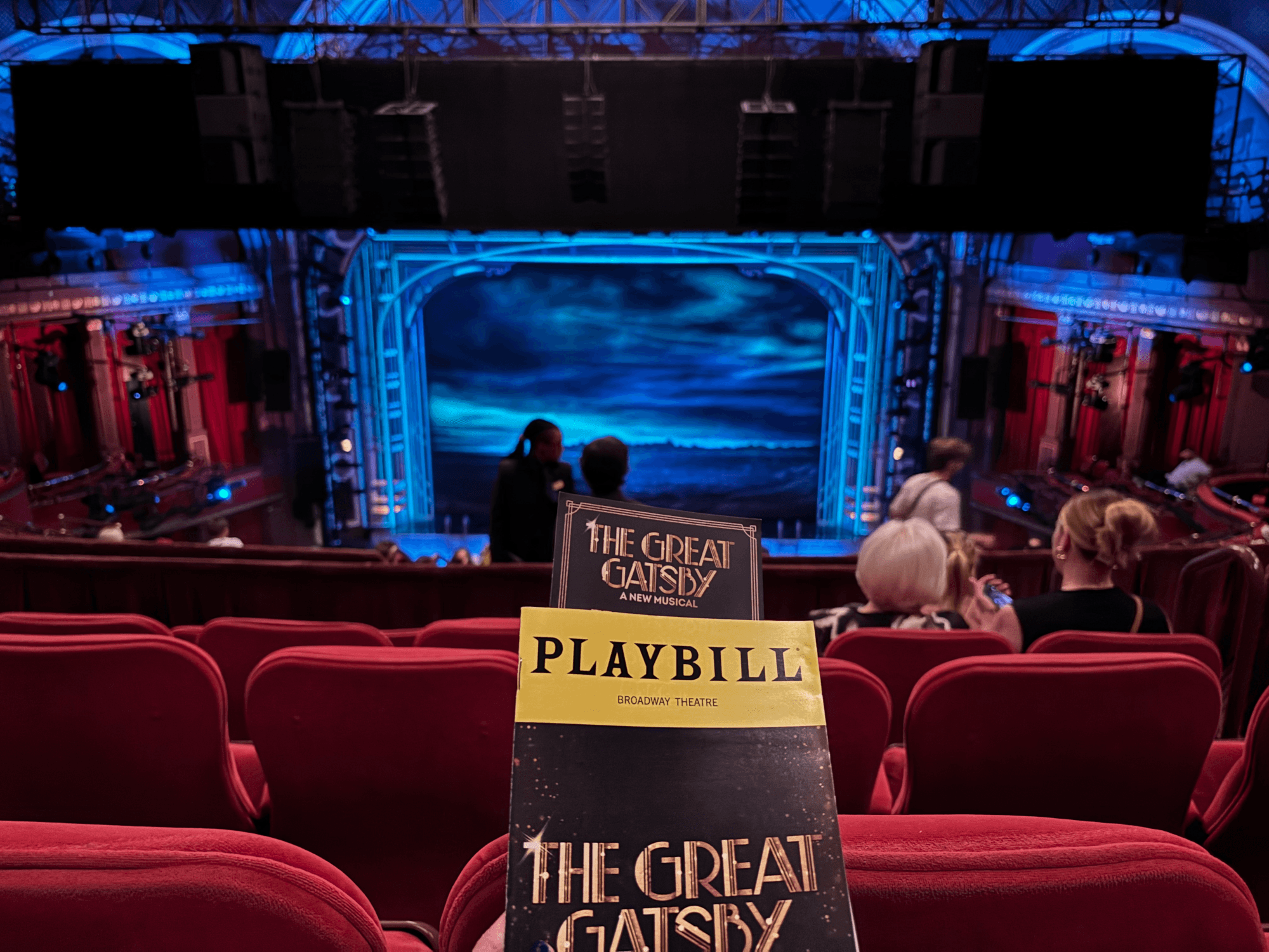 Picture of the Broadway Theater from inside. In the background the stage is illuminated by a blue glow, with the picture of the aurora in the skies in the backdrop. The theater seats are empty in front of me, and the playbill of the play is being held in the foreground.