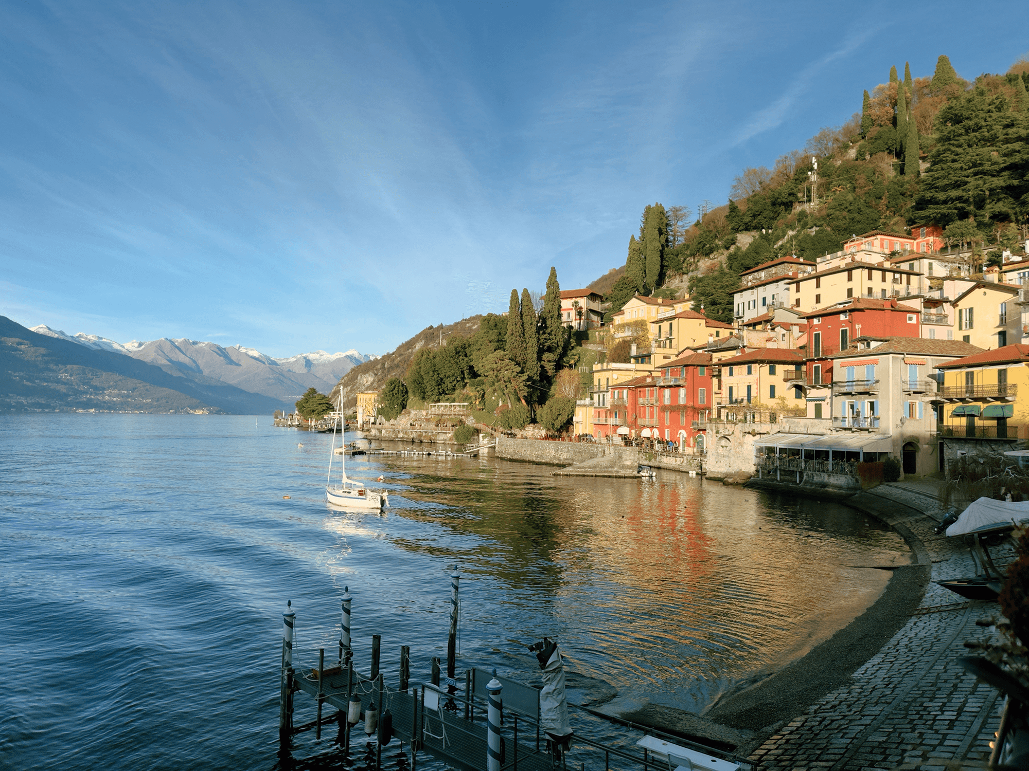 Picturesque medieval-like italian village on the shore of a huge blue lake. The houses are very colorful and there's a lot of pine trees on the slope. A small boat floats on the lake, and in the background sit the snow-capped Alps.