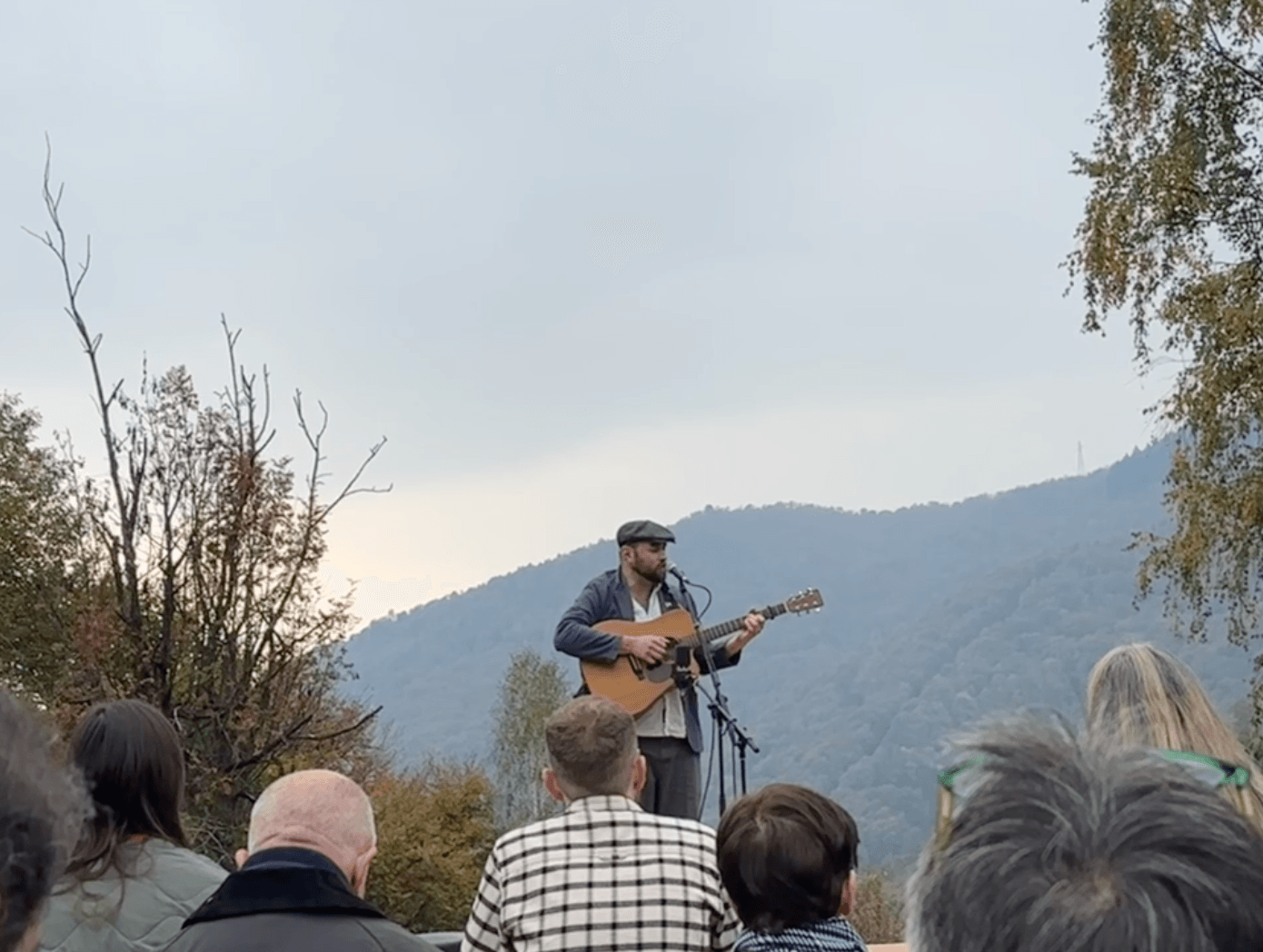 Musician Nick Hart performing with acoustic guitar at outdoor mountain venue, with forested hillside in background and small audience seated below.