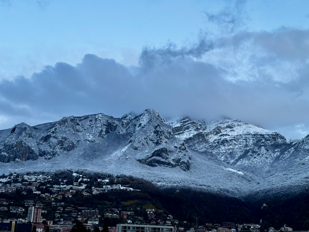 Snowcapped mountains slightly obscured by clouds, with a gray sky. Some houses appear on the foot of the mountains.